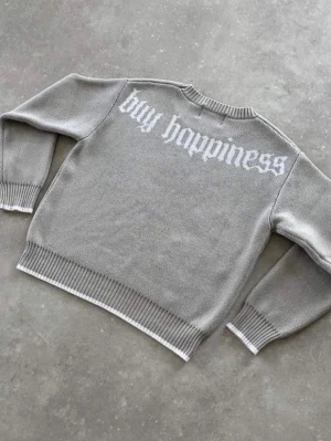 Grå stickad tröja med text - Säljer en grå stickad tröja med vit texten 'buy happiness' på ryggen och en stor dollar-symbol på framsidan. Tröjan har rund hals, ribbade muddar och nederkant samt en avslappnad passform. Perfekt för dig som gillar streetwear och statement-plagg.