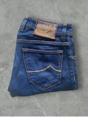 Mörkblå jeans från Jacob Cohen - Snygga mörkblå jeans från Jacob Cohen med klassisk femficksmodell och kontrastsömmar. Jeansen har en läderpatch med logga bak i midjan och raka ben. Tillverkade i mjukt denimtyg som ger en skön passform och stilren look.