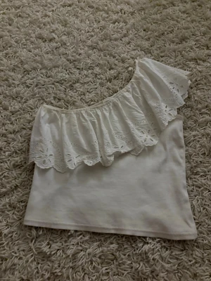 Vit oneshoulder topp med volang Zara - Superfin vit oneshoulder topp från Zara med bred volangkant i broderad spets. Toppen är ribbad och har en snygg asymmetrisk design som ger en trendig look. Perfekt att styla med jeans eller kjol för en cool outfit.