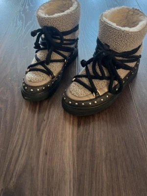 Fodrade beige kängor Inuikii - Säljer ett par beige kängor från Inuikii med mjukt teddyfoder och svarta snören som går runt hela skon. Yttermaterialet är i fårskinn och sulan är svart med nitar runtom. Perfekta för kalla dagar och riktigt snygga till jeans eller leggings.