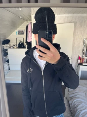 Svart hoodie med dragkedja Les Deux - Säljer en svart hoodie från Les Deux med dragkedja framtill och justerbar huva. Hoodien har en liten vit logga på bröstet och är tillverkad i mjukt bomullsmaterial. Perfekt för en avslappnad och stilren look.