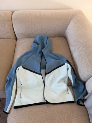 Nike blå och vit hoodie med dragkedja - Snygg hoodie från Nike i ljusblått och vitt med svarta detaljer. Tröjan har dragkedja framtill, huva och Nike-logga på bröstet. Tillverkad i mjukt material som känns skönt mot huden. Perfekt för en sportig och avslappnad stil. 