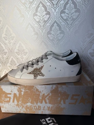 Golden Goose vita sneakers med glitter - Golden Goose sneakers i vitt läder med grå snörning, silverglittrig stjärna på sidan och svart glitterdetalj på hälen. Klassisk låg modell med slitna detaljer för en cool och trendig look. NYPRIS 6000, helt nya använda Max 1 gång inomhus.✅ äktenhetsbevis finns såklart alltid✅🧾