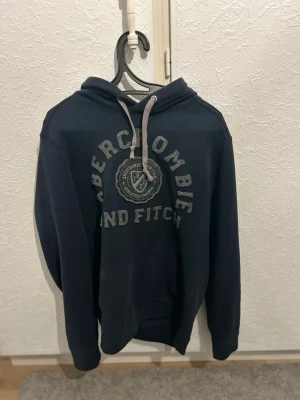 Mörkblå hoodie Abercrombie & Fitch - Snygg mörkblå hoodie från Abercrombie & Fitch med grå dragsko och stor logga broderad på bröstet. Klassisk känguruficka framtill och mjukt material som känns skönt mot huden. Perfekt för en avslappnad och trendig look.