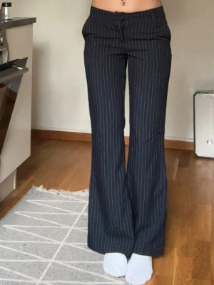 Mörkblå randiga kostymbyxor - Snygga mörkblå kostymbyxor med vita smala ränder och utsvängda ben. Klassisk midja med knapp och dragkedja framtill samt fickor på sidorna. Perfekt för dig som gillar en stilren och trendig look.