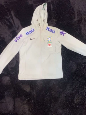 Ny Brazil Vindbreaker – Grön, Storlek M, , Helt Ny utan Prislapp - Upptäck stil och funktionalitet med denna helt nya Brazil vindbreaker i färgen grön, perfekt för alla väder! Tillverkad i 100% polyester, är denna vindjacka både lätt och slitstark, vilket gör den idealisk för både vardagsbruk och sportaktiviteter.  Detaljer: 	•	Färg: Grön 	•	Storlek: M 	•	Material: 100% polyester 	•	Skick: Helt ny, aldrig använd, utan prislapp 	•	Design: Klassisk vindbreaker-stil med elastiska muddar och justerbar huva för extra komfort 	•	Funktion: Vind- och vattenavvisande, p