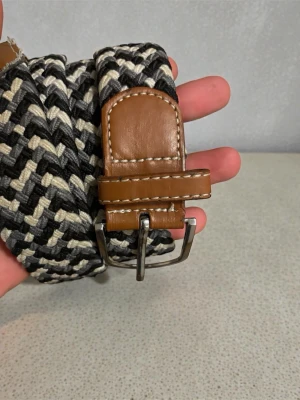 Flätat svartvitt bälte med skinn - Snyggt flätat bälte i svart, vitt och beige med bruna skinn-detaljer vid spännet. Bältet har ett klassiskt metallspänne och passar perfekt till jeans eller chinos. Ger en cool touch till din outfit.