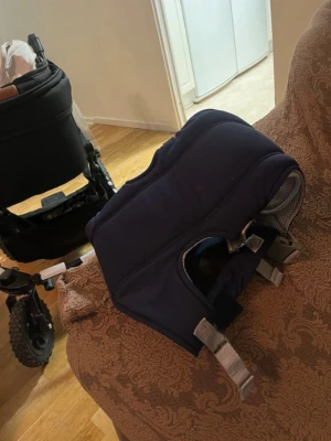 Mörkblå bärsele med justerbara remmar - Säljer en mörkblå bärsele med grå detaljer och justerbara remmar. Selen har vadderade axelband och meshpartier för extra komfort och ventilation. Passar för att bära små barn nära kroppen och är enkel att ta på och av.