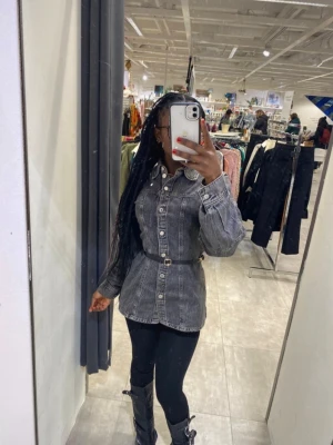 Svartgrå jeansjacka med bälte - Snygg svartgrå jeansjacka med markerad midja och tillhörande bälte. Jackan har klassisk krage, knäppning framtill och långa ärmar. Perfekt för dig som vill ha en trendig och figurnära look. Bältet följer inte med.