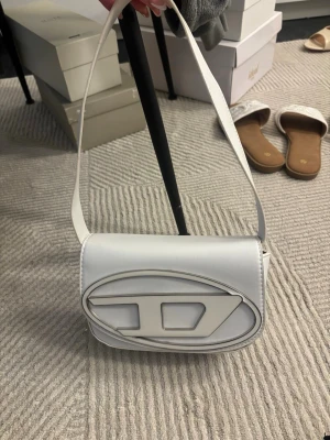 Vit handväska - Liten vit axelväska i stilren design. Slät yta med dekorativ detalj framtill och kort axelrem. Smidig storlek som rymmer det viktigaste. Passar både till vardag och kväll. 