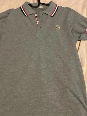 Grå Moncler piké  - Snygg grå pikétröja från Moncler med klassisk krage och knappar framtill. Kragen har stilrena ränder i rött, vitt och blått. Moncler-logga broderad på bröstet. Perfekt till en casual look.