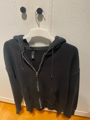 Svart oversized hoodie från H&M - Säljer en svart hoodie från H&M i oversized fit med dragkedja framtill och två stora fickor. Hoodien har justerbar huva med snören och mjukt material på insidan. Perfekt för en chill och avslappnad stil.