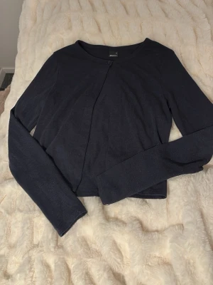 Mörkblå croppad kofta från Gina Tricot  - Snygg mörkblå croppad kofta från Gina Tricot med lång ärm och enkel knappstängning framtill. Perfekt att slänga över en topp för en clean och stilren look. Tunn och mjuk i materialet.