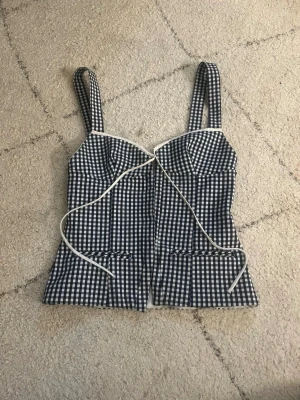 Rutig korsettopp från Zara - Supersnygg svartvit rutig korsettopp från Zara med breda axelband och dekorativ snörning framtill. Toppen har en figurnära passform och vita detaljer längs kanterna. Perfekt för dig som vill sticka ut lite extra i sommar!