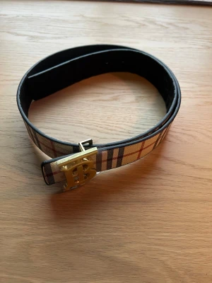 Rutmönstrat Burberry bälte med guldspänne - 105 cm, Snyggt bälte från Burberry med klassiskt rutmönster i beige, svart och rött. Bältet har ett stort guldfärgat spänne med Burberrys ikoniska TB-logga. Perfekt accessoar för att lyfta din outfit.