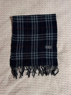 Rutig halsduk från Burberrys i ull - Snygg halsduk från Burberrys i klassiskt rutigt mönster med svarta, gråa och röda toner. Tillverkad i 100% lammull och har fransar i ändarna. Perfekt accessoar för att lyfta din outfit med en tidlös brittisk stil.