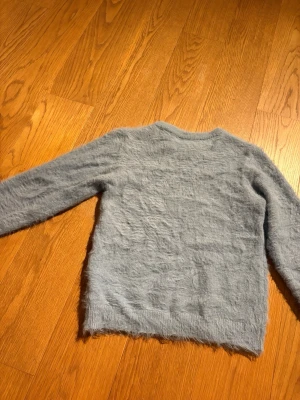 Ljusblå fluffig stickad tröja SAMSA - Mysig ljusblå stickad tröja från SAMSA Young i storlek 170. Tröjan har en fluffig, mjuk yta och klassisk rund hals. Perfekt för dig som gillar en chill och bekväm stil. Långa ärmar och enkel design gör den lätt att matcha.