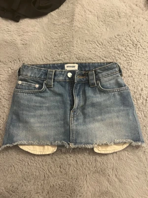 Blå jeanskjol från Weekday - Säljer en kort blå jeanskjol från Weekday med rå kant nertill och klassiska fem fickor. Kjolen har bälteshällor och dragkedja framtill. Perfekt för dig som gillar en avslappnad och trendig stil.