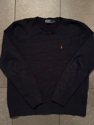 Mörkblå stickad tröja Polo Ralph Lauren - Klassisk mörkblå stickad tröja från Polo Ralph Lauren med rund halsringning och ribbade muddar. Tröjan har den ikoniska röda logotypen broderad på bröstet. Perfekt till jeans eller över en skjorta för en clean look.