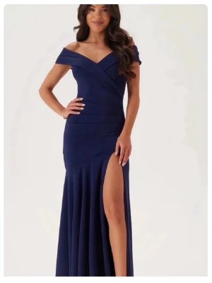 Marinblå långklänning med slits - Elegant marinblå långklänning med offshoulder och v-ringad överdel. Klänningen har en snygg slits framtill och draperade detaljer över byst och midja. Perfekt för bal eller festliga tillfällen. Bara använd 1 gång