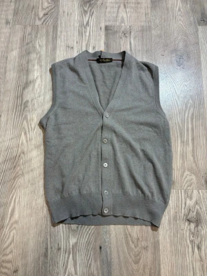 Loro Piana Väst - Klassisk grå stickad väst från Loro Piana med v-ringning och knäppning framtill. Västen har ribbade kanter nedtill och vid ärmöppningarna, vilket ger en snygg och stilren look. Perfekt att bära över en skjorta för en clean outfit. Storlek 54 passar S