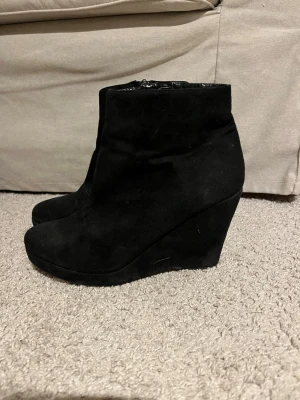 Svarta kilklackar - Säljer ett par svarta stövletter med kilklack och dragkedja på insidan. Skorna är i mockaimitation. Väl använda men med rätt behandling kan de bli fina igen!