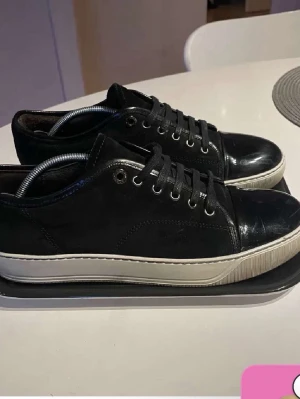 Glans lanvins  - Snygga svarta sneakers med ovandel i mocka och detaljer i blankt skinn på tån och runt snörningen. Skorna har klassisk snörning och vit sula som ger en stilren kontrast. Perfekta för dig som gillar en clean och modern look.