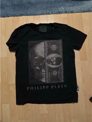 Svart Philipp Plein t-shirt med tryck - Cool svart t-shirt från Philipp Plein med stort tryck av döskalle och sedel på framsidan. Klassisk passform med korta ärmar och rund hals. Märkeslapp i sidan. Perfekt för dig som gillar edgy streetstyle.