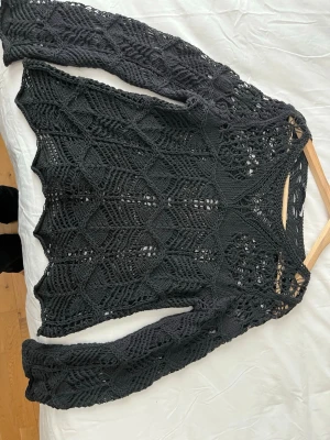 Svart virkad stickad tröja - Säljer en svart stickad tröja med virkat mönster och vida ärmar. Tröjan har ett genombrutet, luftigt mönster och rund halsringning. Perfekt för lager på lager och ger en cool boho-vibe till din outfit.