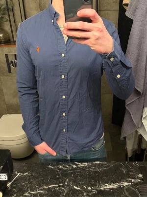 Blå skjorta från Ralph Lauren - Klassisk blå skjorta från Ralph Lauren med button-down krage och broderad orange logga på bröstet. Skjortan har lång ärm, vita knappar och en slim passform. Perfekt till jeans eller chinos för en clean och stilren look.