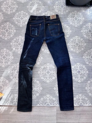 Mörkblå jeans från Nudie Jeans - Snygga mörkblå jeans från Nudie Jeans med klassisk femficksdesign och kontrastsömmar. Jeansen har raka ben och är tillverkade i kraftig denim. Läderpatch med logga baktill i midjan. Perfekta för en avslappnad och trendig look.