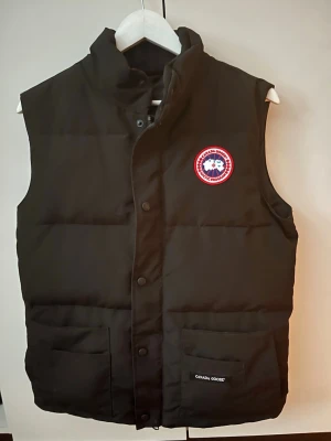 Canada Goose väst - Stilren svart dunväst från Canada Goose med hög krage, två stora fickor framtill och klassisk logotyp på bröstet. Västen har tryckknappar och dragkedja samt är vadderad för extra värme. Perfekt för lager-på-lager och streetstyle.