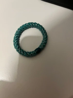 Turkos glittrig hårsnodd - En elastisk hårsnodd i turkos med glittrig finish. Snodden är rund och har en liten plastdetalj som håller ihop ändarna. Perfekt för att sätta upp håret med en snygg touch av färg och glitter.