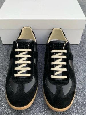 Maison Margiela sneakers - Stilrena svarta sneakers från Maison Margiela med ovandel i mocka och läder, vita snören och beige innersula. Klassisk låg modell med gummisula och diskret logga på plösen. Perfekta för dig som gillar minimalistisk och tidlös design.