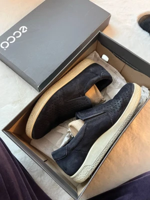 Mörkblå loafers från ECCO i mocka - Säljer ett par mörkblå loafers från ECCO i mjuk mocka med perforerade detaljer på ovandelen. Skorna har en vit, mönstrad sula och är slip-on-modell för enkel användning. Perfekta för en stilren och avslappnad look.