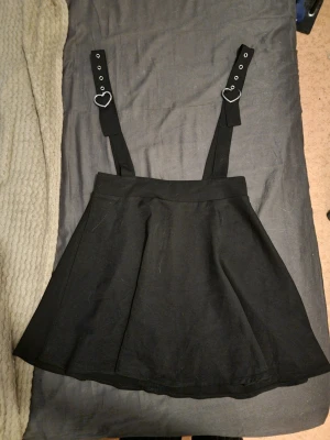 Svart kjol med hängslen och hjärtdetaljer - Säljer en svart kjol med breda hängslen som har silverfärgade hjärtformade spännen. Kjolen har en utsvängd form och är gjord i ett mjukt tyg, troligen bomull eller polyester. Perfekt för en edgy och söt stil.