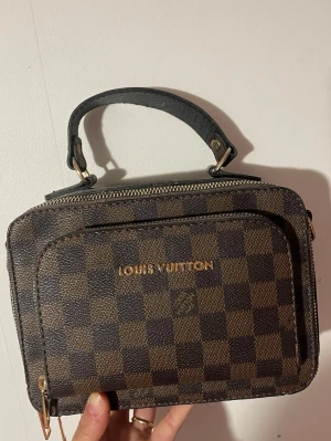 Damier handväska brun - Snygg handväska i klassiskt brunt Damier-mönster. Väskan har ett topphandtag, guldiga metalldetaljer och dragkedja. Tillverkad i canvas med läderdetaljer och har en praktisk ytterficka. Otroligt snygg nu inför våren och sommaren. 