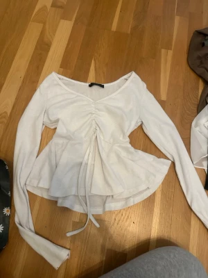 Vit långärmad topp med snörning - Supersöt vit långärmad topp med v-ringning och snörning framtill som ger en snygg rynkad effekt. Toppen har volang nertill och är gjord i ribbad bomull. 