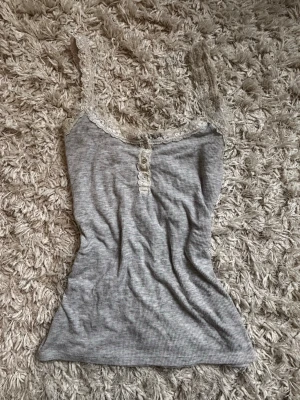 Grå ribbad spetslinne Hunkemöller - Grå ribbad linnetopp från Hunkemöller med fina spetsdetaljer runt urringningen och smala axelband. Toppen har knappar framtill och är gjord i ett mjukt, stretchigt material som passar perfekt för mysiga hemmakvällar.