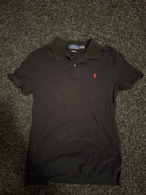 Svart pikétröja från Polo Ralph Lauren - Klassisk svart pikétröja från Polo Ralph Lauren med korta ärmar och krage. Tröjan har den ikoniska röda logotypen broderad på bröstet och är gjord i mjuk bomull. Perfekt till jeans eller shorts för en clean och stilren look.