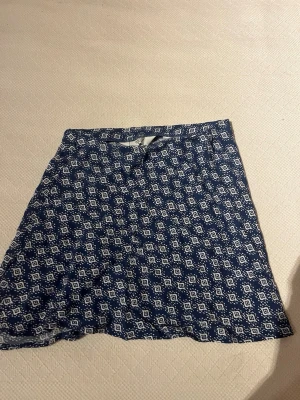 Blåvit mönstrad kortkjol från H&M - Kortkjol från H&M i blått med vitt geometriskt mönster. Kjolen har en A-linjeformad siluett och är tillverkad i ett mjukt bomullsmaterial som känns skönt mot huden. Perfekt för dig som gillar en avslappnad men ändå trendig stil.