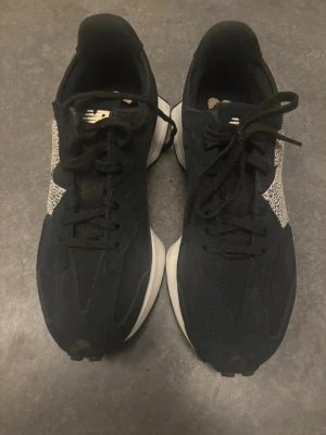 Svarta New Balance sneakers med mönstrad N-logga - Snygga svarta sneakers från New Balance med en stor vit N-logga på sidan som har ett coolt svartprickigt mönster. Skorna har svarta snören, svart ovandel i textil och mocka samt en vit chunky sula. Perfekta för dig som gillar streetstyle och vill sticka ut. Använda en gång