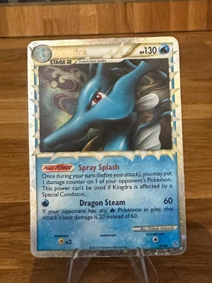 Kingdra Pokémonkort holo - Säljer ett holografiskt Kingdra Pokémonkort med blå och silver färgtema. Kortet har 130 HP och visar Kingdra med attackerna Spray Splash och Dragon Steam. Kortet är från 2010 och har en glittrig ram och detaljerad illustration.