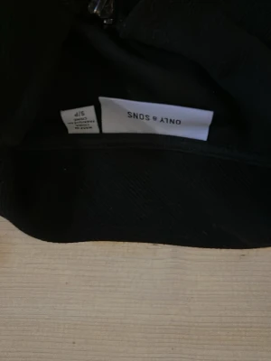Svart topp från ONLY & SONS - En stilren svart topp från ONLY & SONS med enkel design. Toppen har korta ärmar och rak passform, perfekt för en clean och modern look. Materialet känns mjukt och följsamt mot huden.