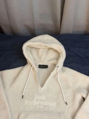 Vit teddyhoodie från Peak Performance sälj inte längre!!!  - Mysig vit teddyhoodie från Peak Performance med broderad logga på bröstet. Hoodien har dragkedja framtill, justerbar huva med snören och en stor magficka. Tillverkad i fluffigt teddy-material som håller dig varm och bekväm