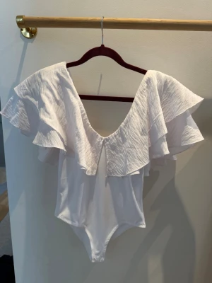 Vit body med volang från Zara - Supersnygg vit body från Zara med djup urringning både fram och bak. Stora dubbla volanger över axlarna ger en trendig och romantisk look. Tillverkad i ett lätt, stretchigt material som sitter bekvämt på kroppen. 