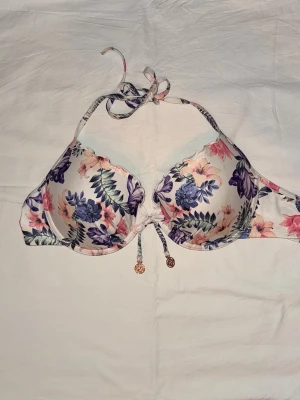 Bikinitopp  - Snygg bikiniöverdel med blommigt mönster i lila, rosa och grönt på vit botten. Modellen har vadderade kupor, halterneck-knyt och dekorativ snörning framtill med små guldfärgade detaljer. Går att justera storleken lite på bandet runt om finns 3 lägen så den går att få mindre runt om Perfekt för strandhäng eller poolparty! 