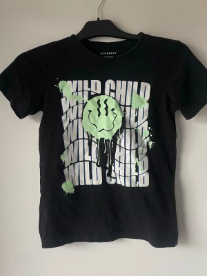 Svart t-shirt Wild Child Firefly size 134-140 - Cool svart t-shirt från Firefly med trycket 'WILD CHILD' i vitt och en grön smiley med färgstänk på framsidan. T-shirten har rund hals och korta ärmar, perfekt för en avslappnad och edgy stil. Storlek 134-140