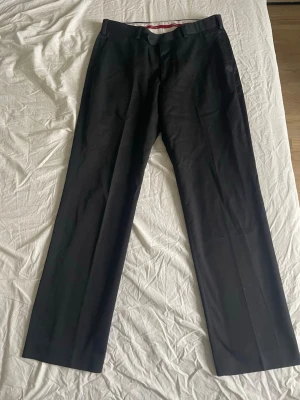 Svarta kostymbyxor med raka ben - Snygga svarta kostymbyxor med klassisk rak passform och pressveck. Byxorna har hällor för skärp och två bakfickor med knapp. Perfekta till en dressad look och passar till skjorta eller kavaj.