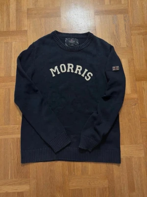 Morris mörkblå stickad tröja - Mörkblå stickad tröja från Morris med vit broderad logotyp på bröstet och Union Jack-detalj på ärmen. Rund halsringning och ribbade muddar vid ärmslut och nederkant. Perfekt för dig som gillar klassisk och stilren design. Står storlek Large men den är liten i storleken och passar M mycket bättre 